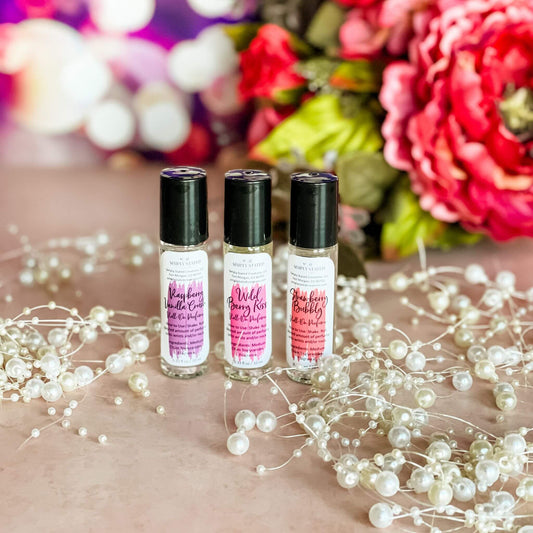 A trio of Sweet Temptations Roll-On Perfumes—Raspberry Vanilla Crush, Strawberry Bubbly, and Wild Berry Kiss—arranged on a blush pink background with pearl accents and vibrant floral decor.